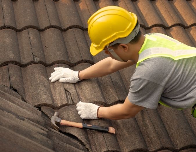 Man wearing a hard hat on top of a roof repairing shingles.